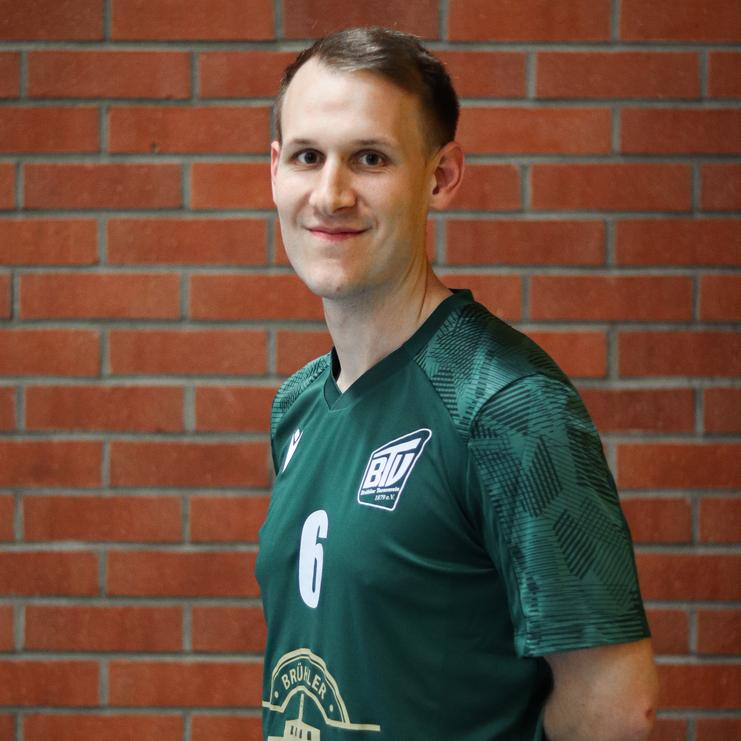 Porträt eines Mannes in einem grünen Sporttrikot mit der Nummer 6, lächelnd vor einer Backsteinwand.