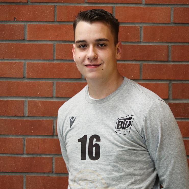 Junger Mann in Grauem Sweatshirt mit der Nummer 16 steht vor einer roten Ziegelwand und lächelt in die Kamera.