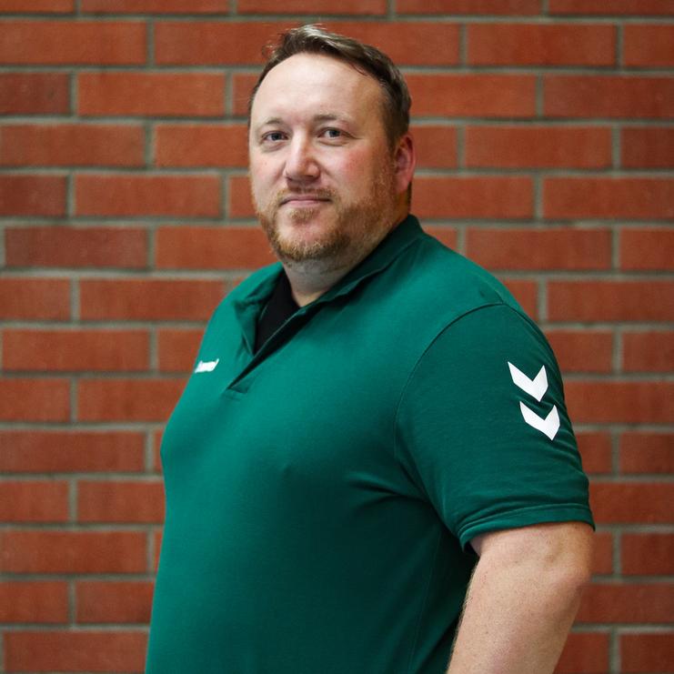 Porträt eines Mannes in grünem Polohemd, lächelnd vor einer Ziegelwand, mit einer Hand auf der Hüfte.