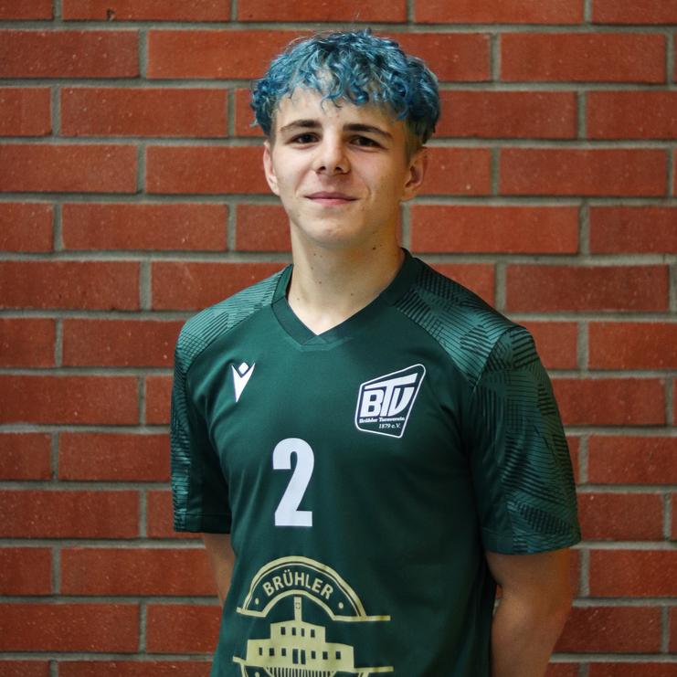 Junger Mann mit blauen Locken in grünem Fußballtrikot vor einer roten Mauer, Nummer 2 auf dem Trikot.