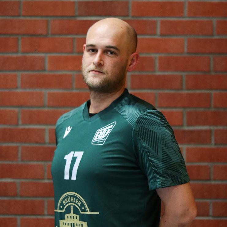 Mann in grüner Sporttrikots mit der Nummer 17 steht vor einer roten Ziegelwand und blickt zur Seite.