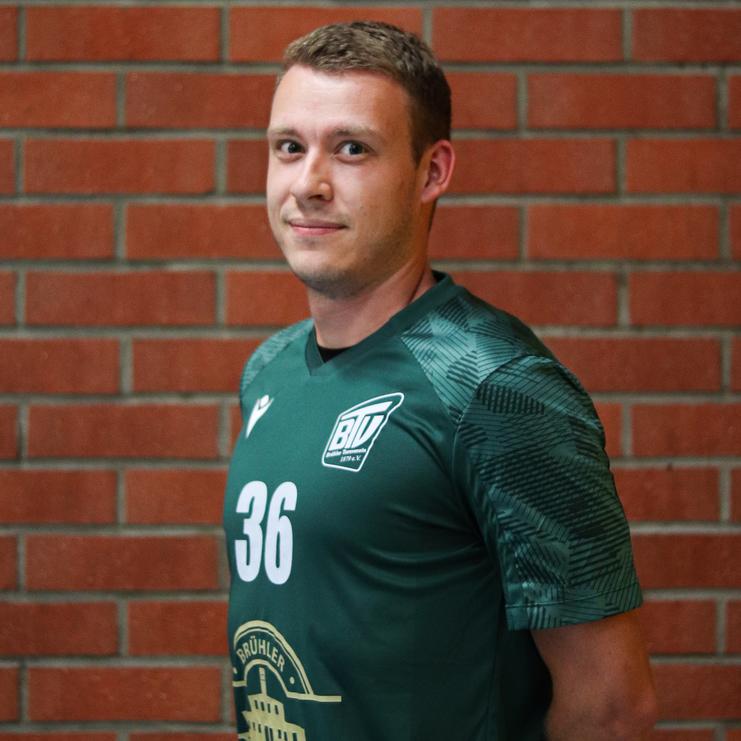 Porträt eines lächelnden Mannes in grünem Sporttrikot mit der Nummer 36, vor einer Backsteinwand stehend.