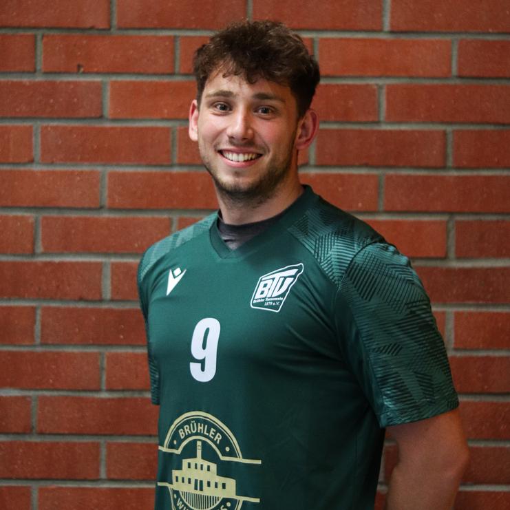 Lächelnder junger Mann in grünem Sporttrikot mit der Nummer 9, vor einer Backsteinwand stehend.