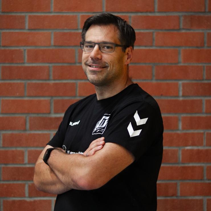 Mann mit Brille und schwarzem Shirt steht an einer Backsteinwand, die Arme vor der Brust verschränkt und lächelt.