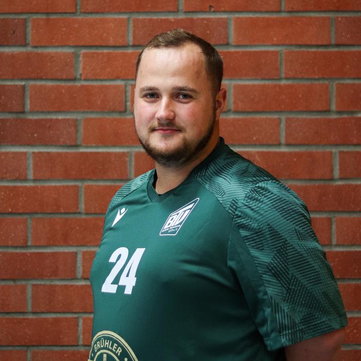 Mann in grüner Sporttrikot mit der Nummer 24 steht vor einer Ziegelsteinmauer und schaut in die Kamera.