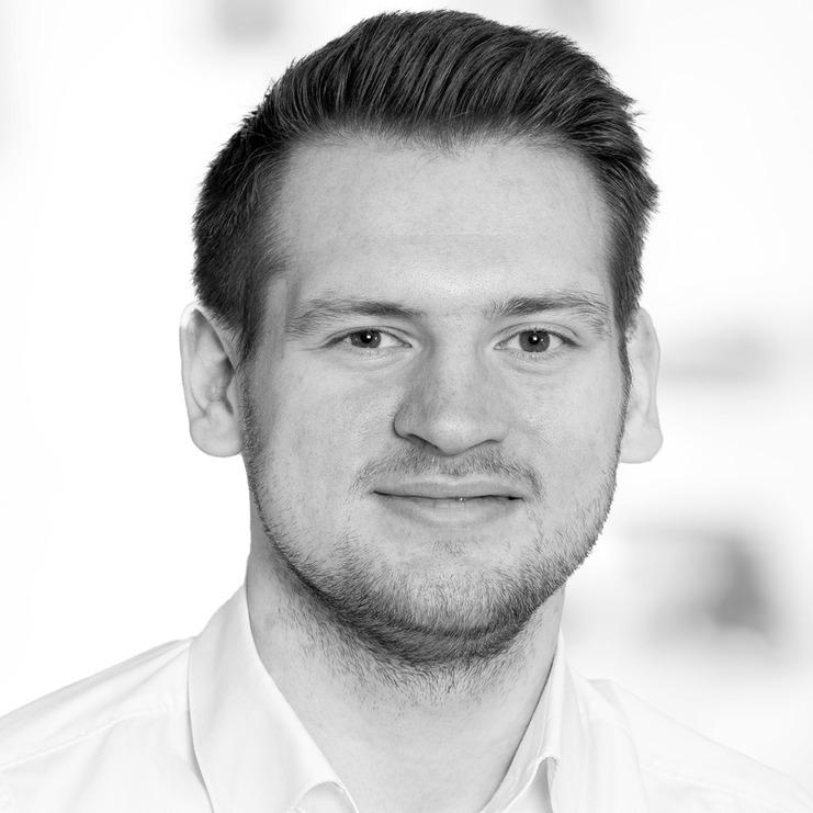 Schwarz-weiß-Porträt eines lächelnden Mannes mit kurzen Haaren, der ein weißes Hemd trägt, vor unscharfem Hintergrund.