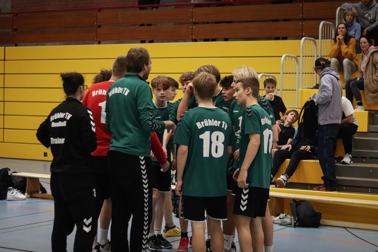 Die Handballmannschaft Brühler TV bespricht sich vor einem Spiel in einer Sporthalle. Zuschauer sitzen im Hintergrund.