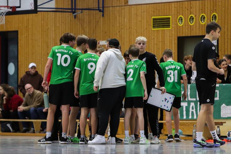 Gruppe von Handballspielern in grünen Trikots versammelt sich an der Seitenlinie, während ein Trainer Anweisungen gibt.