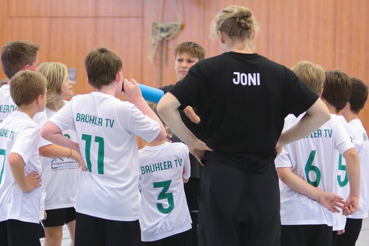 Gruppe von Jungen in weißen T-Shirts mit „BRÜHLER TV“ versammelt um einen Trainer in schwarzem Shirt in einer Sporthalle.