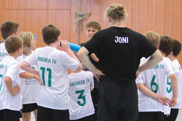 Gruppe von Jungen in weißen T-Shirts mit „BRÜHLER TV“ versammelt um einen Trainer in schwarzem Shirt in einer Sporthalle.