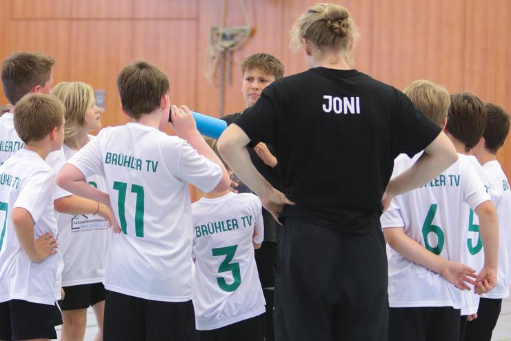Gruppe von Jungen in weißen T-Shirts mit „BRÜHLER TV“ versammelt um einen Trainer in schwarzem Shirt in einer Sporthalle.