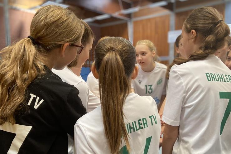 Gruppe junger Mädchen in Sporttrikots bespricht sich in einer Sporthalle. Nummern und Vereinsnamen auf den Shirts sichtbar.