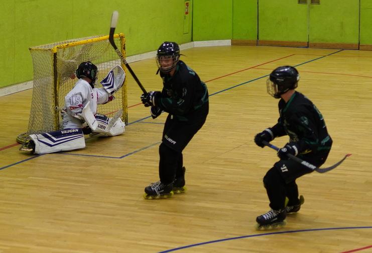 Zwei Rollhockeyspieler in schwarzer Ausrüstung vor einem Torwart in weißer Kleidung auf einem Sportfeld.