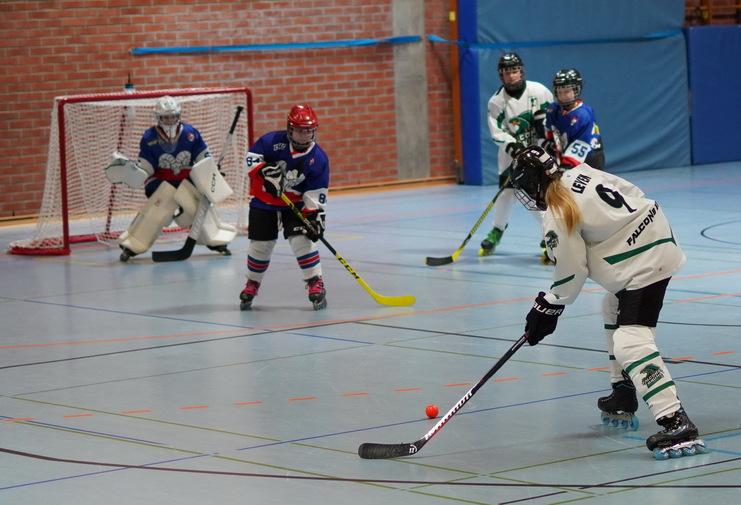 Fünf Spielerinnen beim Inline-Hockey, eine schießt auf das Tor, während die Torhüterin bereitsteht.