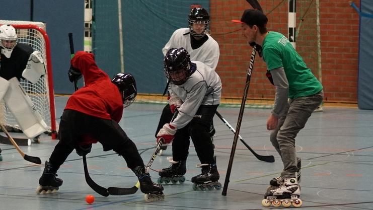 Sportliche Szene mit Spielern in Rollschuhen, die beim Inline-Hockey um den Ball kämpfen, vor einem Tor.