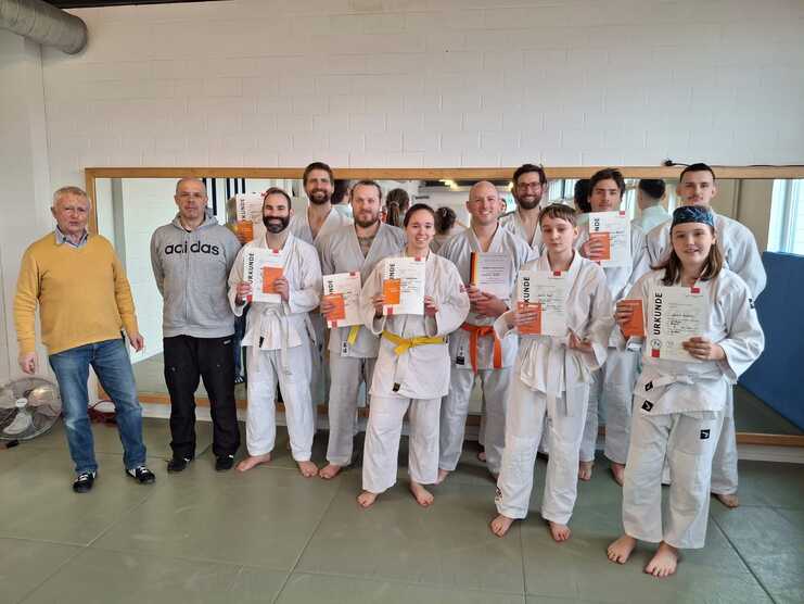Gruppe von elf Personen in Ju-Jutsuanzügen steht barfuß in einer Trainingshalle vor einem Spiegel und hält Urkunden in die Kamera. Zwei Männer in Alltagskleidung stehen links daneben. Die Gruppe wirkt zufrieden und stolz, nach einer b