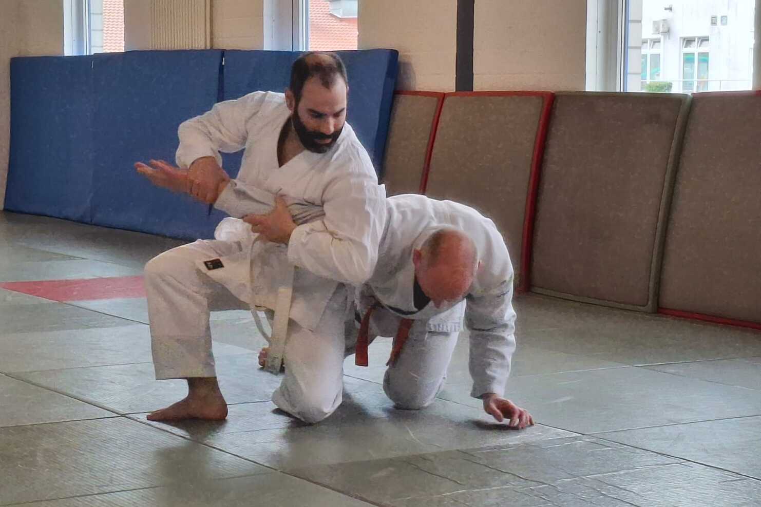 Zwei Personen in Karateanzügen trainieren in einer Halle auf Matten: Eine Person fixiert den Arm der anderen in einer Hebeltechnik, während diese kniend nach vorne gebeugt ist. Im Hintergrund stehen gepolsterte Matten an der Wand