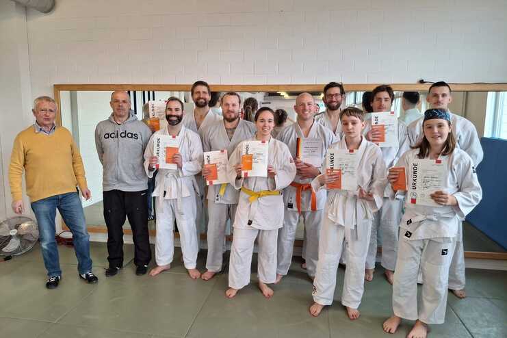 Gruppe von elf Personen in Ju-Jutsuanz&uuml;gen steht barfu&szlig; in einer Trainingshalle vor einem Spiegel und h&auml;lt Urkunden in die Kamera. Zwei M&auml;nner in Alltagskleidung stehen links daneben. Die Gruppe wirkt zufrieden und stolz, nach einer b