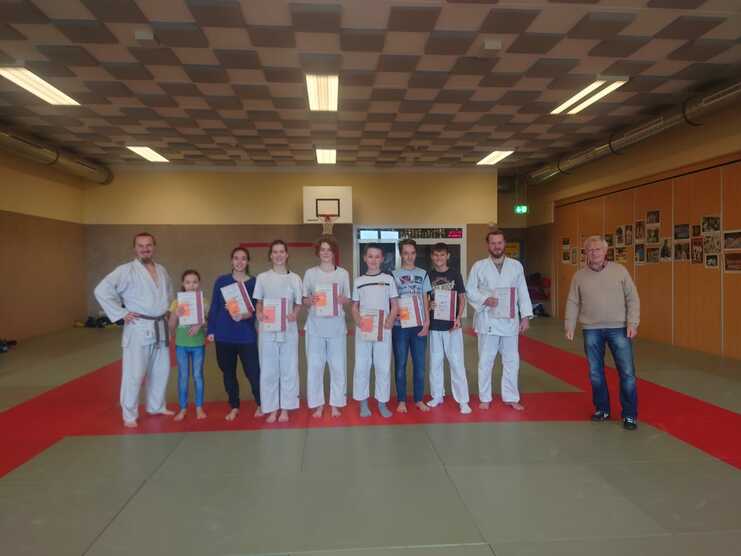 Gruppe von fünf Jugendlichen und zwei Erwachsenen im Judogi hält Urkunden auf einer roten Judomatte in einem Trainingsraum.