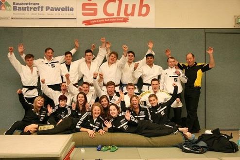 Gruppe von Judokas in Uniformen, die freudig posieren, auf Matten und einer Sportbank, mit erhobenen Händen.