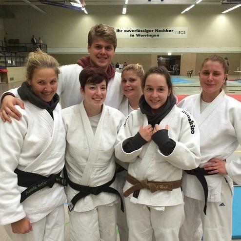 Sechs Judokas in weißen Gi-Anzügen posieren lachend für ein Gruppenfoto in einer Sporthalle.