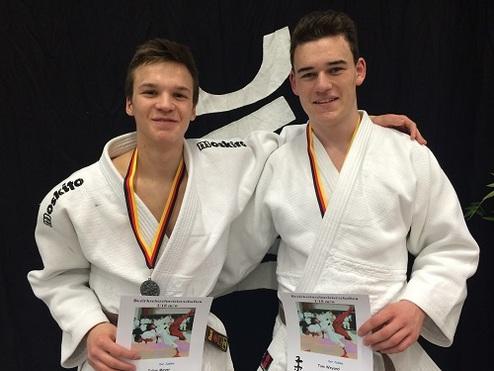Zwei Judoka in weißen Gi mit Medaillen halten Urkunden in der Hand, vor einem dunklen Hintergrund.