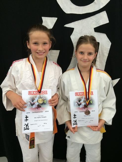 Zwei Mädchen in Judogi halten Urkunden und Medaillen nach einem Wettkampf vor einem schwarzen Hintergrund.