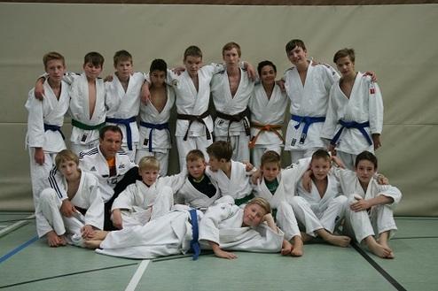 Gruppe von 16 Jugendlichen in Judoanzügen, posierend mit einem Trainer in einer Turnhalle.
