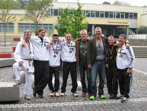 Gruppe von sieben Männern in Sportkleidung mit Medaillen, posierend vor einer Schule im Freien.