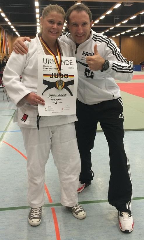 Junge Frau in Judo-Gi hält Urkunde mit ihrem Namen; daneben steht ein Trainer, beide lächeln.