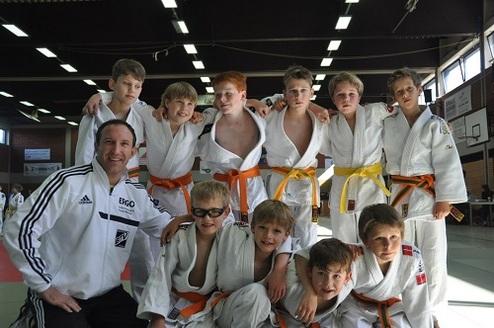 Gruppe von zehn Judokas in weißen und orangen Judogi lächelt auf einer Wettkampfbahn, ein Trainer steht vorne.