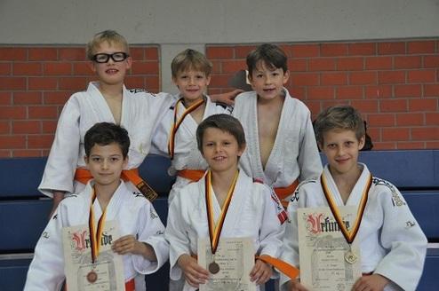 Sechs Kinder in Judoanzügen posieren stolz mit Medaillen und Urkunden in einem Sportumfeld.