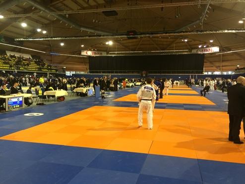 Judo-Wettkampf in einer Halle mit orange-blauen Matten, Zuschauern und Kämpfern in Judogis.