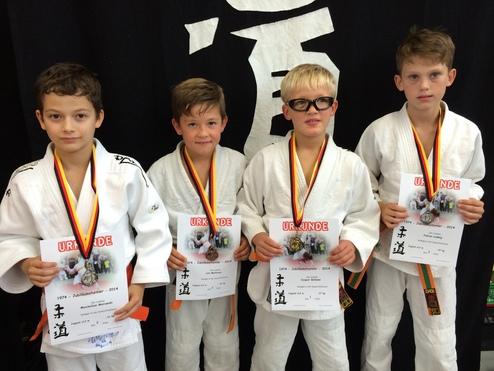 Vier Kinder in Judoanzügen halten Urkunden und Medaillen vor einem schwarzen Hintergrund mit dem Schriftzeichen „Judo“.