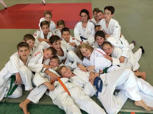 Gruppe von Kindern in Judokleidung, die zusammen auf einer Matte liegen und lächeln. Judostreifen in verschiedenen Farben sichtbar.