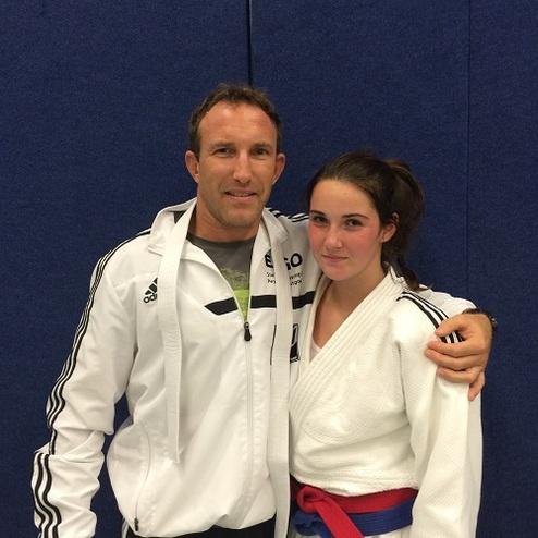 Ein Trainer und eine Judoka posieren lächelnd nebeneinander, beide in Sportkleidung, vor einer blauen Wand.