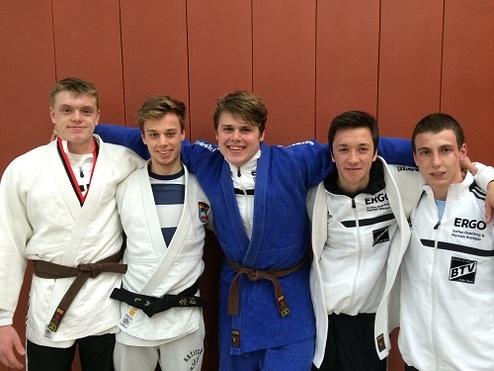 Fünf Männer in Judogi stehen lächelnd nebeneinander vor einer roten Wand.