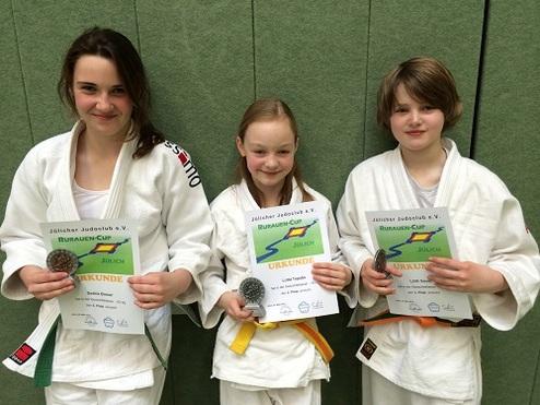 Drei Kinder in Judokleidung halten Urkunden des Rubin-Cups, jeder mit einer Medaille um den Hals, vor einer grünen Wand.