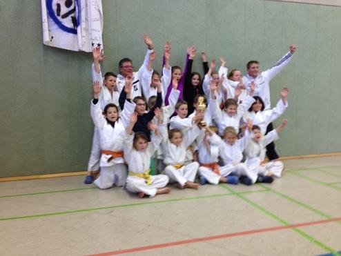 Gruppe von Kindern in Judoanzügen feiert einen Sieg mit einem Pokal in einer Sporthalle.