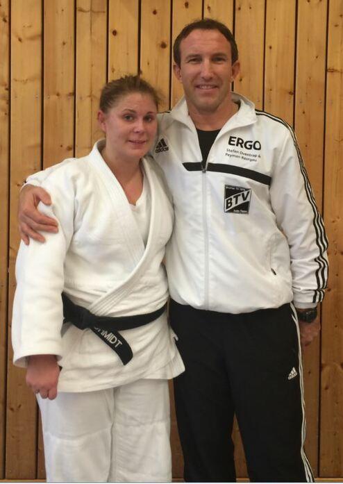 Zwei Judoka posieren lächelnd vor einer Holzwand, die Frau trägt einen weißen und der Mann einen schwarzen Judoanzug.