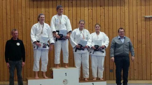 Vier Medaillenträgerinnen im Judo stehen auf dem Siegertreppchen, umrahmt von zwei Personen in Sportkleidung.