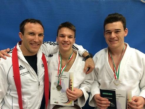 Drei Judokas posieren lächelnd mit Medaillen und Urkunden vor einem blauen Hintergrund.