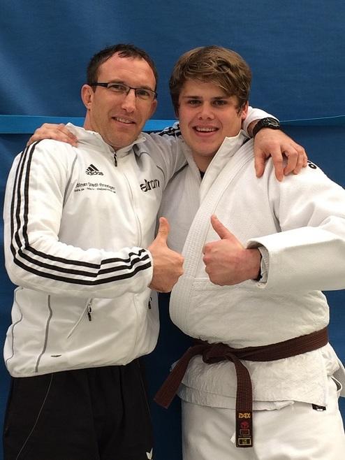 Zwei Judo-Trainer posieren lächelnd mit Daumen-hoch vor einem blauen Hintergrund. Der jüngere trägt einen weißen Judoanzug.