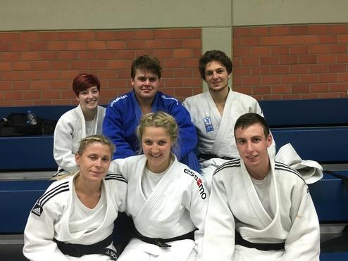 Sechs Judo-Praktizierende in weißen und blauen Judogis sitzen auf einer Tribüne in einer Sporthalle.