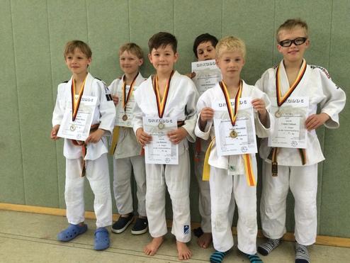Sechs Kinder im Judogi mit Medaillen und Urkunden posieren stolz vor einer grünen Wand in einem Sportraum.