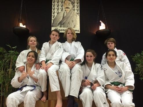 Sechs Jugendliche in weißen Judo-Anzügen sitzen auf einer Holzbank vor einer Wand mit einem Porträt und Fackeln.