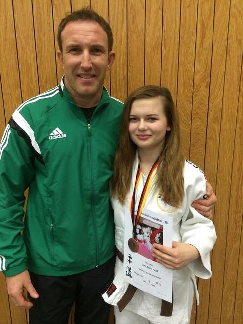 Junge Judoka mit Medaille und Urkunde posiert neben Trainer in Sportjacke vor einer Holzverkleidung.