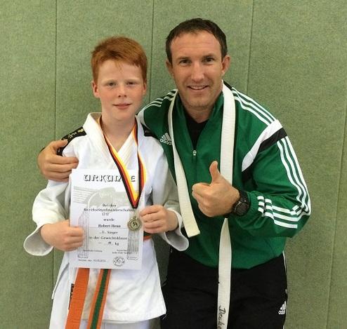 Junge mit Urkunde und Medaille steht neben Trainer, beide lächeln vor grünem Wandhintergrund.