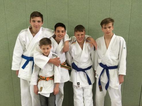 Fünf Jungen in Judogis posieren lächelnd vor einer grünen Wand, einige mit verschiedenen Gürtelfarben.