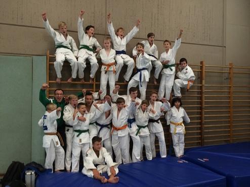 Gruppenzusammenhalt von Judokas in weißen Anzügen mit farbigen Gürtel, die fröhlich auf einer Trainingsmatte posieren.